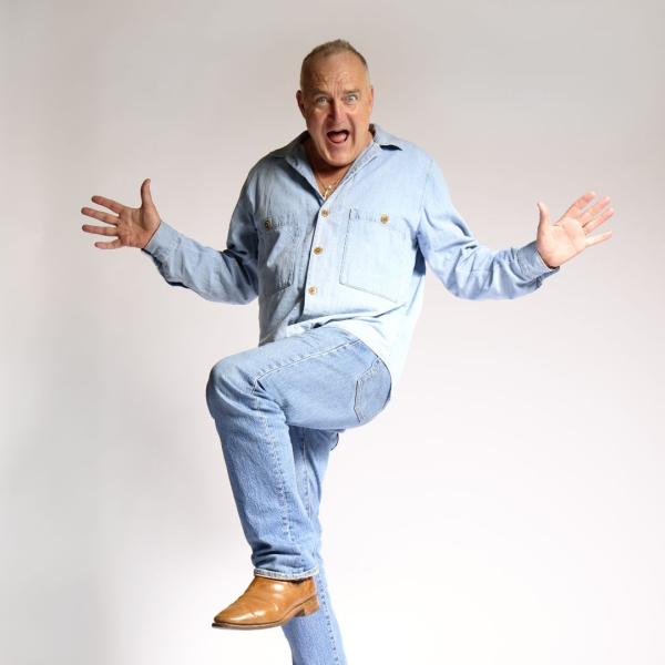 Wearing a light blue shirt and jeans, Jimeoin poses on one leg with his arms outstretched and mouth open.