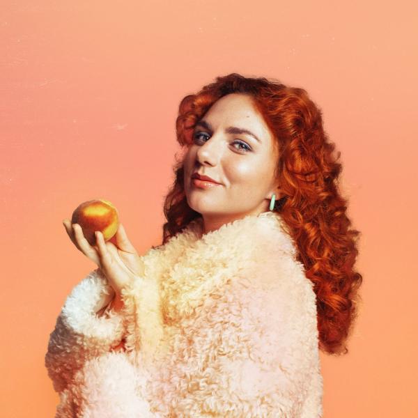 Ania Magliano stands in profile, wearing a fluffy coat and holding a peach in her hand. She smiles against a plain peach background.