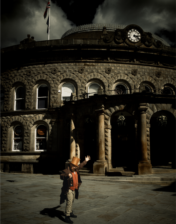 A man wearing an orange top hat and victorian clothing stands holding a lantern with an arm outstretched in the air in front of Leeds Corn Exchange