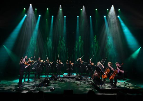 The image shows a group of musicians on a dimly lit stage, arranged in a semicircle and playing string instruments under dramatic green and white spotlights. Cables, music stands, and microphones surround the performers, creating the setup for a live orchestral performance.