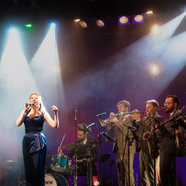 From left to right, a stage is filled with suited musicians: a drummer, two trumpet players and a saxophonist. In the foreground is a glamorous female vocalist below a hazy spotlight.