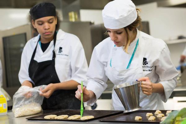 Two female culinary students in a commercial kitchen are focused on preparing pastries on baking sheets. Both wear white chef coats with a Leeds City College logo and head coverings, one in a white cap and the other in a black cap with a black apron. The student in the foreground uses a green pastry brush to glaze small, piped dough rings, while the other student works with a bag of ingredients in the background. The setting features professional stainless steel surfaces and kitchen equipment.