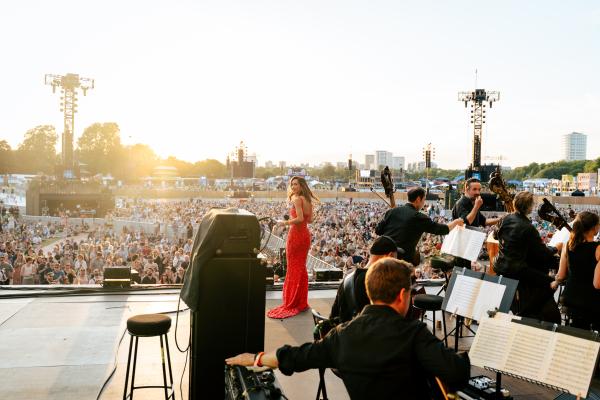 an orchestra performing on a large outdoor stage to a large crowd on a sunny day 