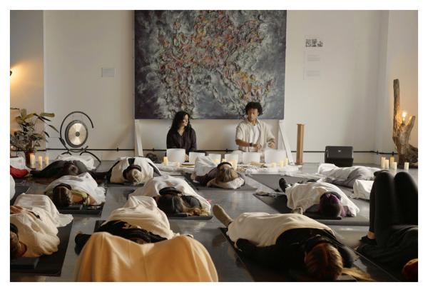 Two people leading a sound bath in a white room with a large canvas above their heads. Participants are laid down on yoga mats and are covered with white blankets and eye masks.