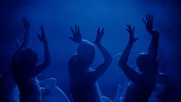 A blue image with the figures of performers dancing on stage in a circle with their arms in the air above them. 