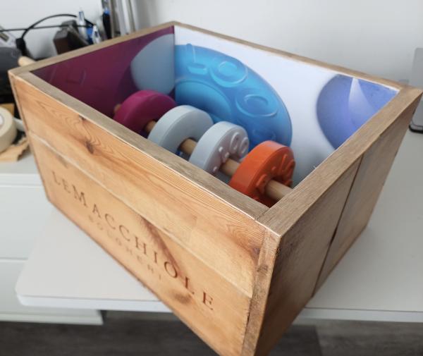 A wooden crate, open at the top to show its contents: four different coloured rings threaded onto a pole fixed across the box’s centre