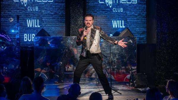 A white man with dark hair and a moustache poses with a microphone against a blue brick background.