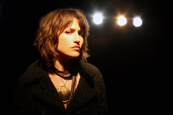 Joan As Police Woman looking directly at the camera, wearing a black textured coat and gold necklace, set against a dark background with three warm stage lights.
