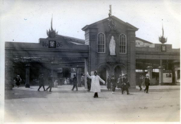 An image of Leeds City Square in 1926, with model moot hall erected for the tercentenary celebrations of Leeds getting its charter. A large building facade with a white statue in a central alcove (Queen Anne) stands in the centre. On either side the dates 1626 and 1926 are etched in what looks like stone. This is entrance to a station concourse. Pedestrians are milling about in period 1920s dress. A policeman in a white coat and gloves stands beneath the arch directing people.