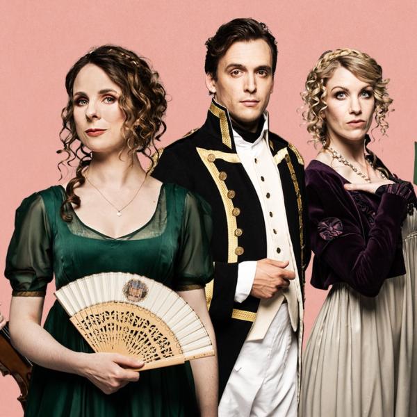 Three people in regal period dresses and a dinner suit stand in front of a plain pink backdrop.