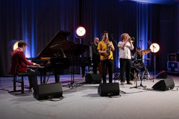 Five musicians playing instruments on an illuminated stage: piano, saxophone, trumpet, drums and double bass.