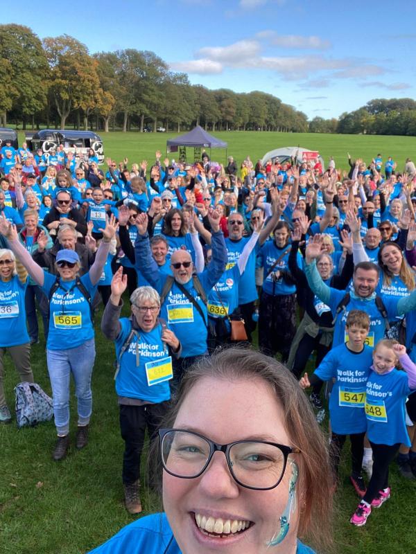 A selfie of Parkinson's UK Community Fundraiser, Ailsa Dragan stood in front of a large group of people all wearing Walk for Parkinson's t-shirts with their arms in the air in celebration