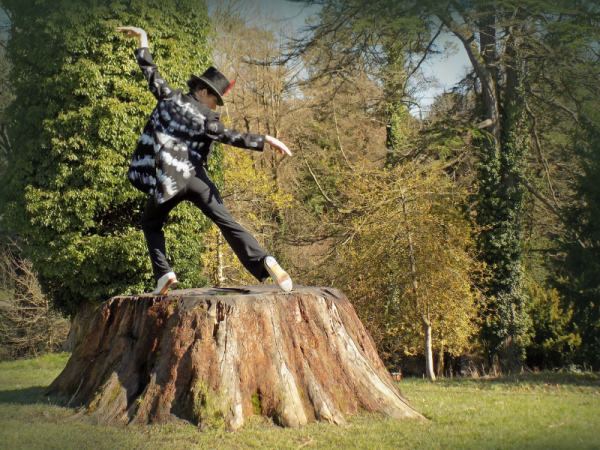 A dancer poses on an extremely large tree stump outdoors in tap shoes, a top hat and tails