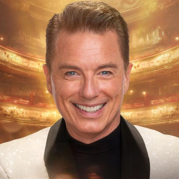 John Barrowman smiles in a sparkling suit in a shimmering auditorium. 