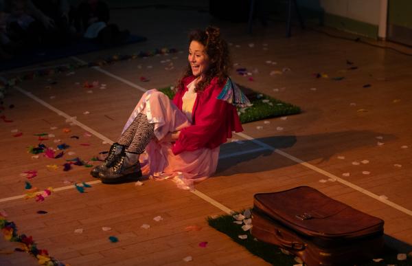 A person sat on a gymnasium floor surrounded by confetti and a suitcase. 