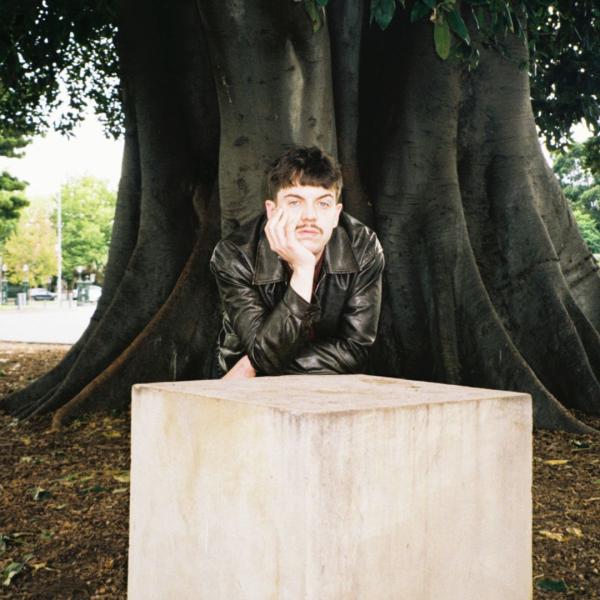A man in a leather jacket rests his head in his palm, his arms folded across a white concrete block in a forest.
