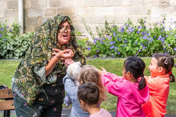 A woman wearing a green shawl and glasses acts shocked in front of a group of smiling children