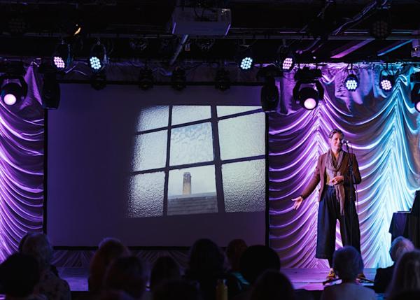 Artist Hannah Lamb standing on the stage of the Old Woollen, with purple and blue lights glowing against a ruched backdrop. She is standing in front of a microphone stand whilst gesturing toward a projector showing an image of a Mill chimney through an old window.
