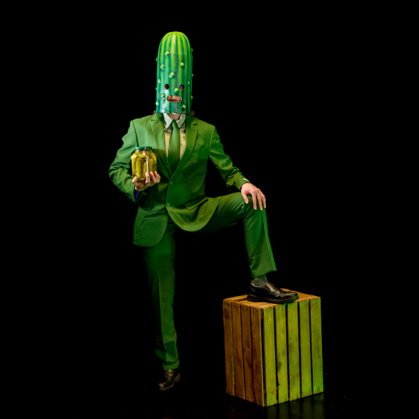 Someone in a green suit, with a cucumber mask holding a jar of gherkins