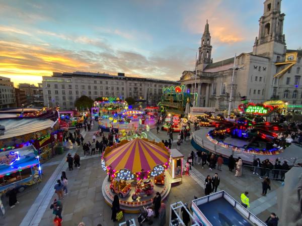 A wide view of a brightly lit funfair at sunset in a city square, with colourful rides, food stalls, and crowds of visitors. 