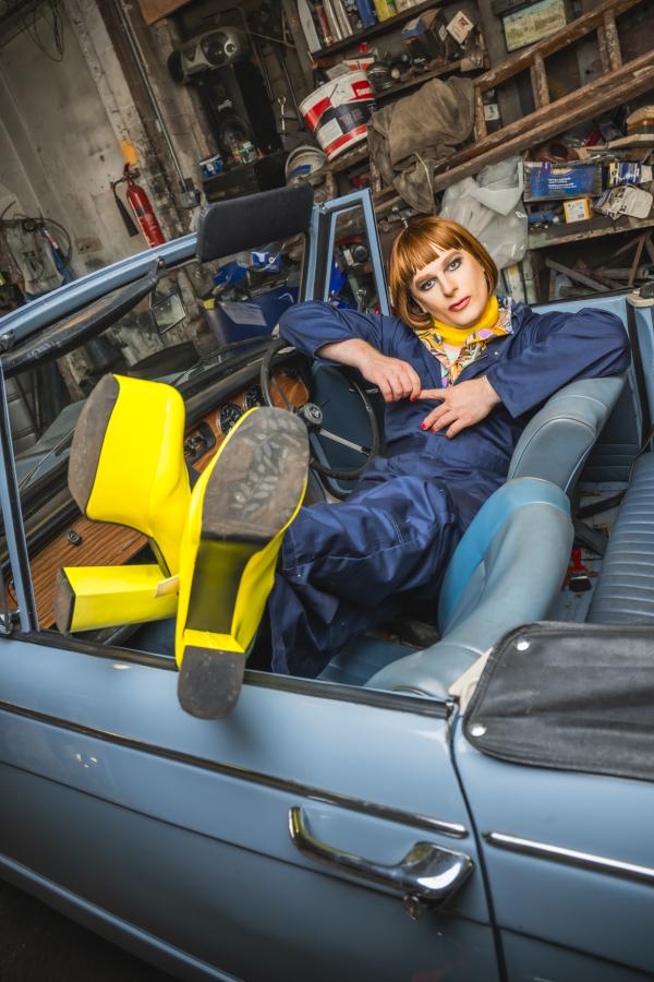 Someone in blue overalls and yellow high heels slouches in a light blue convertible car