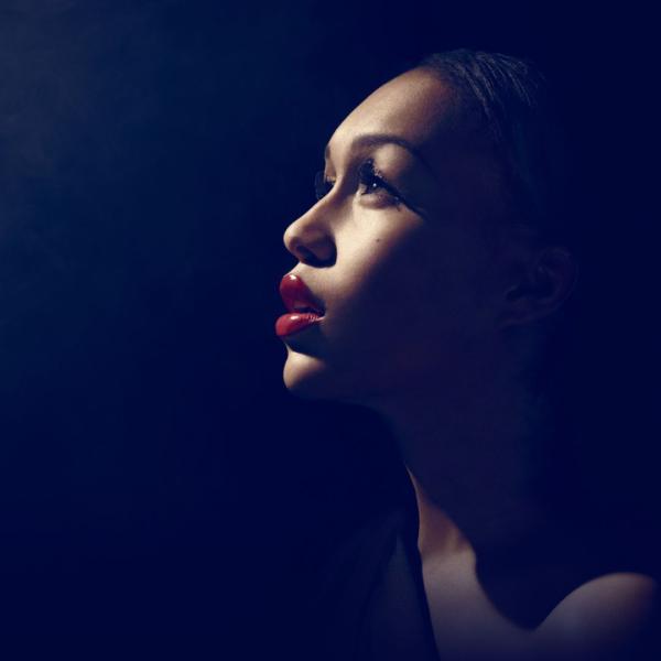 Lit by a solitary spotlight against a dark, hazy backdrop, Rebecca Ferguson looks into the distance with elegant makeup and red lipstick.