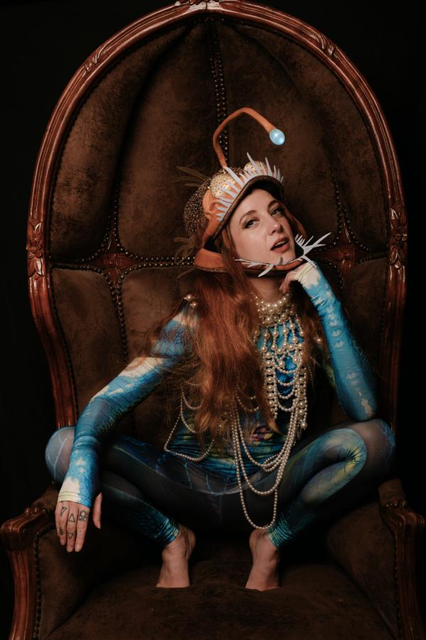Dramatic portrait of a person in a bioluminescent-inspired anglerfish costume, adorned with pearls, seated in a carved wooden throne.