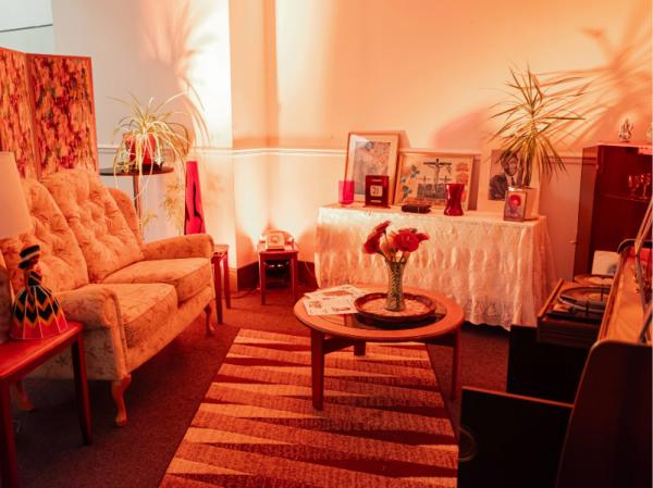 A warmly lit vintage-style living room featuring a floral upholstered sofa, striped area rug, round wooden coffee table with red roses in a vase, and eclectic décor including framed artwork, indoor plants, and a record player. The room is bathed in orange-red ambient lighting, creating a cozy, nostalgic atmosphere with artistic and retro accents throughout.