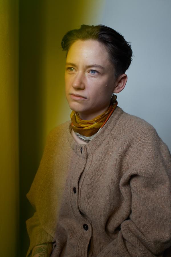 Portrait of a person with short hair wearing a tan cardigan and scarf, featuring dramatic yellow lighting and a contemplative expression