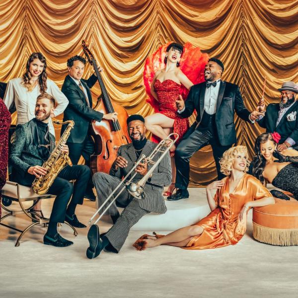 Large ensemble photo of Scott Bradlee's Postmodern Jukebox of a variety of musicians posing with their instruments or candidly smiling in front of a gold curtain, all wearing glamorous ball gowns and vintage suits.