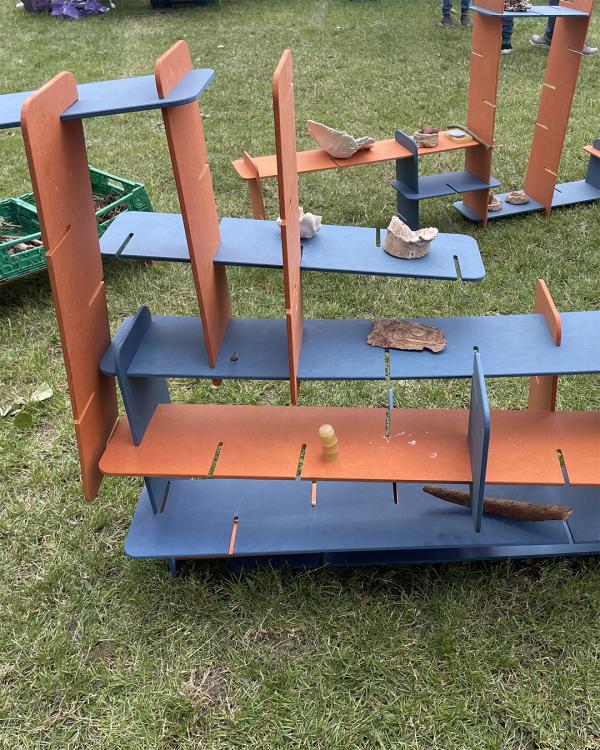 Colorful play structures with orange and blue panels are arranged on grass. Nature items like bark and stones are placed on them. A playful, creative scene.