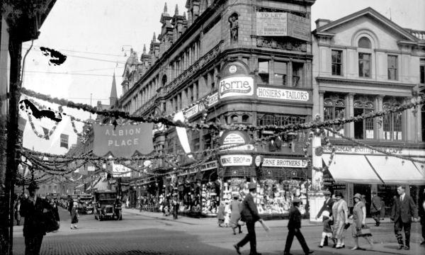 Black-and-white photograph of a busy city street lined with multi-storey shops, decorated with bunting and banners. Pedestrians cross the road while cars and trams travel through the junction. A banner reading “ALBION PLACE” hangs above the street.