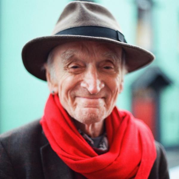Martin Carthy smiles in a brown trilby hat and red scarf, a blue building behind him.