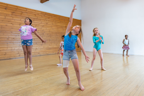 A group of young children are doing ballet in a dance studio, jumping, stretching and swaying.
