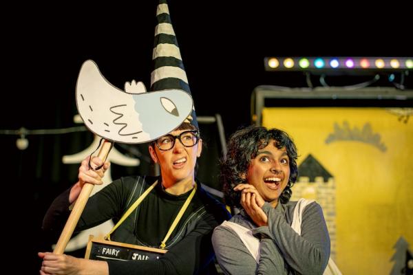 A woman dressed as a dunce holding a stick puppet bird and a girl in a pinafore looking joyful