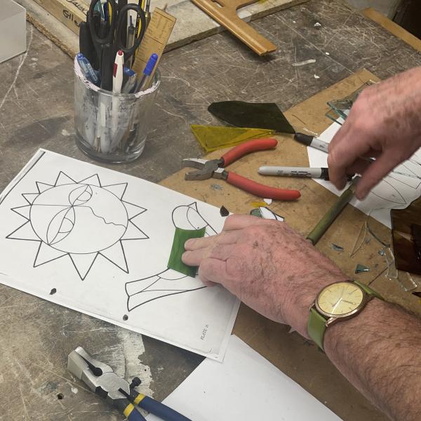 A mans hands holding a piece of green glass on top of a paper template of a bird. 