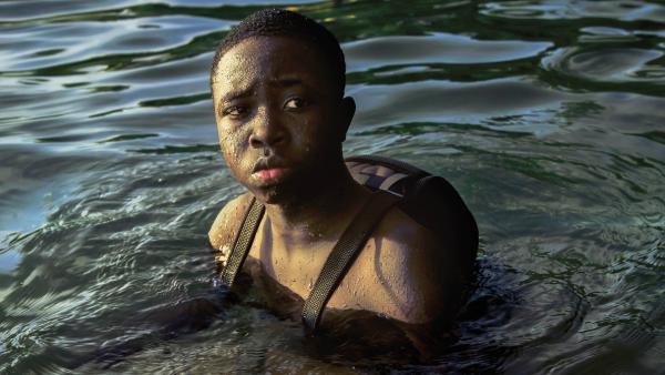 A Black boy with short hair is in water with a backpack 