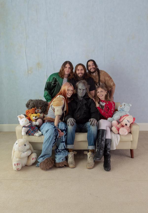 A group of six people cheerfully pose on a sofa surrounded by teddy bears against a light blue backdrop. The central figure, dressed as a goblin, is surrounded by smiling friends.