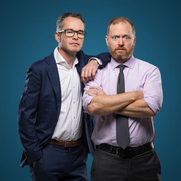 Two men in suits and office attire stand straight-faced against a blue background. One has his arms crossed, the other rests his arm on his shoulder.