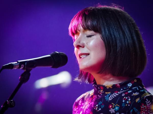 Close up of Emily performing, focused on her. Light shining on her headm eyes cloased singing into a microphone