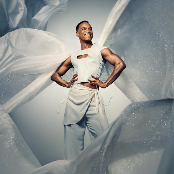 With hands on hips, Johannes Radebe smiles and looks off to the side in a white outfit, surrounded by flowing sheets of sparkling fabric.