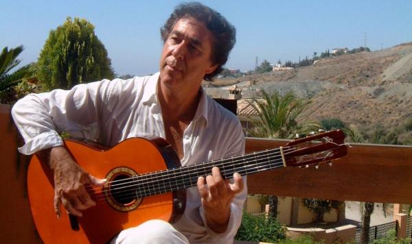 A flamenco guitarist sits, guitar upon his knee, hands held over the strings in that readily identifiable Flemenco style. He is sat in front of a rural Spanish landscape.