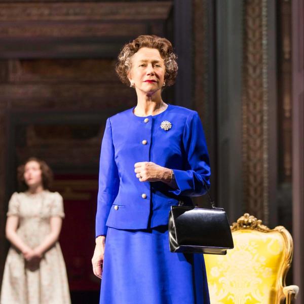 Helen Mirren as Queen Elizabeth II, standing poised on stage in a blue dress and blazer, holding a handbag.