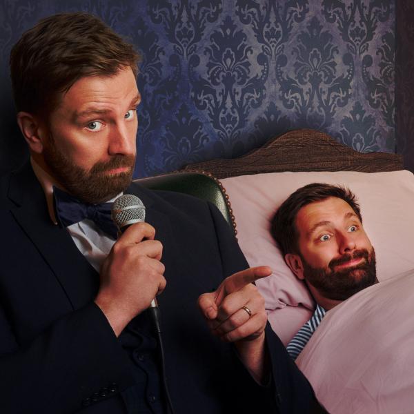 Pierre Novellie sits beside a bed, holding a microphone. In the bed, a second Pierre Novellie is tucked in beneath a pink blanket with a comical smile.