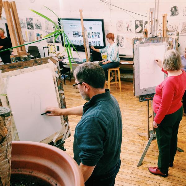 A photograph of three people stood at easels drawing, in the foreground is a man with a green sweater drawing a life model with charcoal