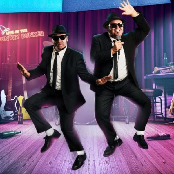 The two suited and shades-wearing Blues Brothers bounce around a neon-lit stage in a dive bar setting featuring guitars and a saxophone.