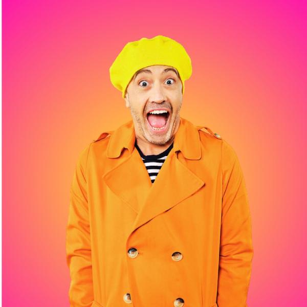 Paulus the Cabaret Geek stands in a bright orange raincoat and yellow hat with a larger-than-life grin, wide-eyed and open-mouthed. The background is a soft pink and orange hue.