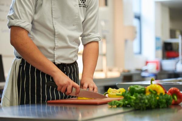 A trainee chef in a white uniform and striped apron slices a yellow bell pepper on a red cutting board in a professional kitchen.
