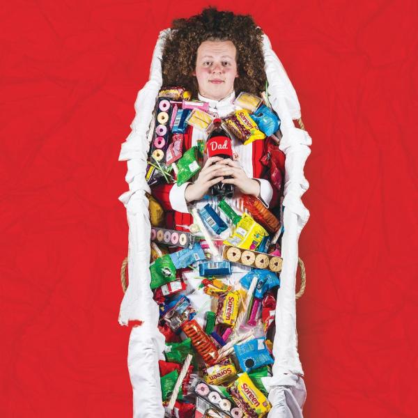 Jack Rooke lies in a coffin-shaped basket full of sweets and biscuits, holding a cola bottle with a label reading 'Dad', surrounded by red material.