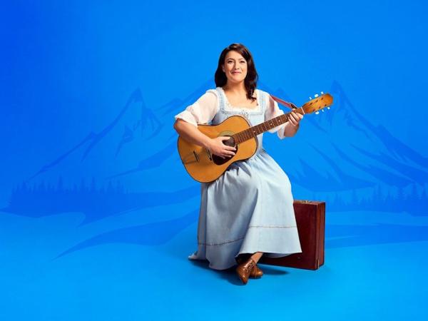 A woman in a light blue dress with long dark hair, sat on a suitcase holding a guitar in front of a blue background of drawn abstract mountain shapes.